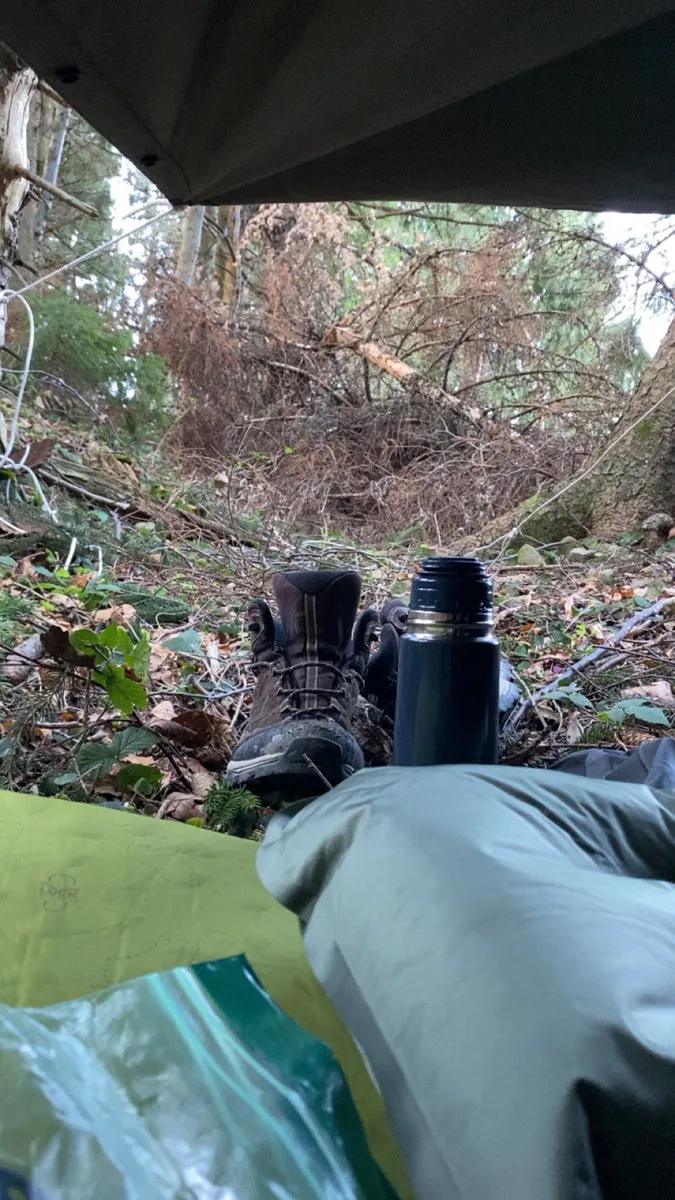 Blick unter einer Plane hinaus auf Wanderschuhe und eine Thermosflasche im Wald